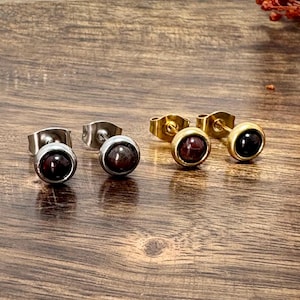 Könnte beinhalten: Eine Auswahl an Ohrsteckern mit runden, dunkelroten und schwarzen Edelsteinen. Die Ohrringe sind in silber- und goldfarbenem Metall gefasst. Der Schmuck wird auf einer Holzoberfläche präsentiert, mit einigen herbstlichen Dekorationen im Hintergrund.