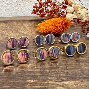 Könnte beinhalten: Eine Sammlung runder Ohrstecker und Ringe mit Holzmaserung in Rosa, Blau und Grau. Die Ohrringe und Ringe haben silber- und goldfarbene Metalleinfassungen. Der Schmuck wird auf einer Holzoberfläche präsentiert.