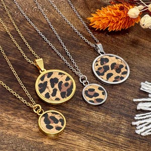 Könnte beinhalten: Vier Halsketten mit runden Anhängern mit Leopardenmuster. Zwei Halsketten haben goldfarbene Ketten und Einfassungen, während die anderen beiden silberfarbene Ketten und Einfassungen haben. Die Anhänger sind mit einem korkartigen Material gefüllt.