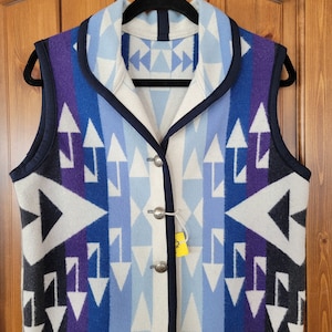 Könnte beinhalten: Eine weiße, blaue, violette und schwarze gemusterte Weste mit Schalkragen und silbernen Knöpfen. Die Weste ist aus einem weichen, gewebten Stoff gefertigt.