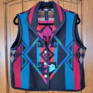 May include: A black and blue patterned vest with a shawl collar and button closure. The vest has a geometric design with red, pink, and green accents. The tag on the vest reads "Scribe Medium".