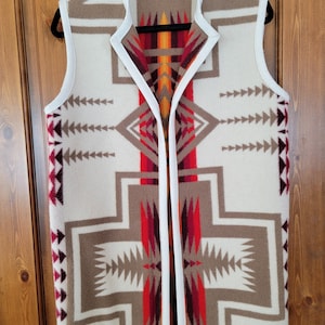 May include: A white sleeveless vest with a geometric pattern in shades of brown, red, and orange. The vest has a white trim around the neckline and armholes.
