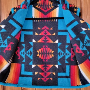 Op de afbeelding: Een turquoise, zwart, rood, oranje en wit gepatroneerd fleecevest met een geometrisch ontwerp. Het vest is opengeknoopt en hangt aan een zwarte hanger.