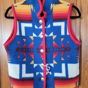 Puede incluir: Un chaleco de vellón azul, rojo, amarillo y blanco con ribete rojo y cierre de botón. El chaleco tiene un diseño del suroeste.