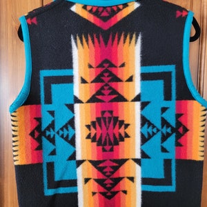 Op de afbeelding: Een zwart, mouwloos vest met een geometrisch patroon in turquoise, rood, oranje en wit. Het vest heeft een ritssluiting en een turquoise rand.