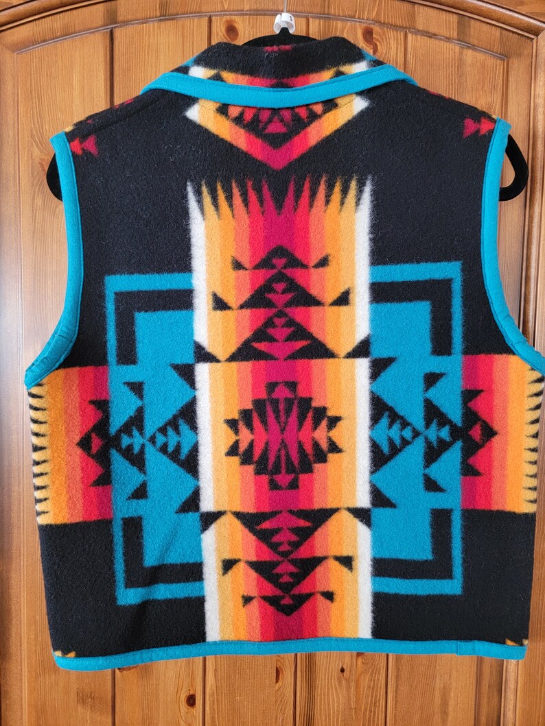 Op de afbeelding: Een zwart, mouwloos vest met een geometrisch patroon in turquoise, rood, oranje en wit. Het vest heeft een ritssluiting en een turquoise rand.