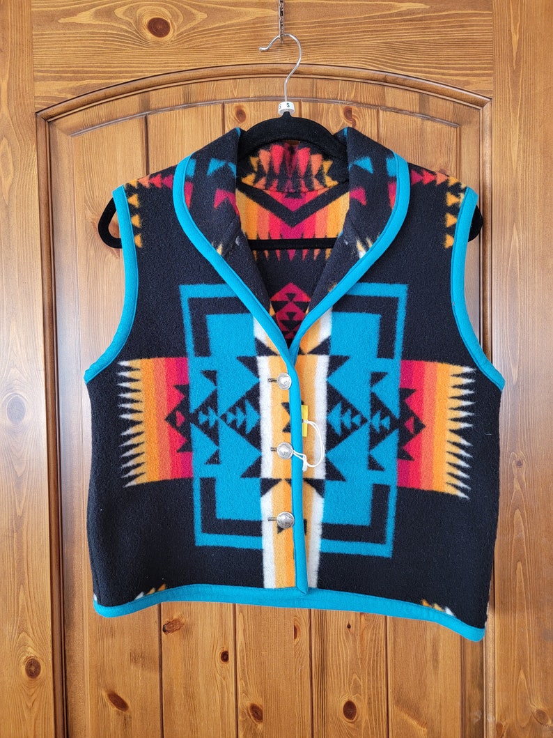 Op de afbeelding: Een zwart fleecevest met een turquoise rand en een geometrisch patroon in zuidwestelijke stijl in tinten oranje, rood, wit en blauw.