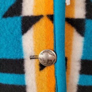 Op de afbeelding: Close-up van een turquoise, zwart, wit en oranje gepatroneerd stof met twee zilveren knopen met een buffelontwerp. De knopen zijn met wit garen aan de stof bevestigd.
