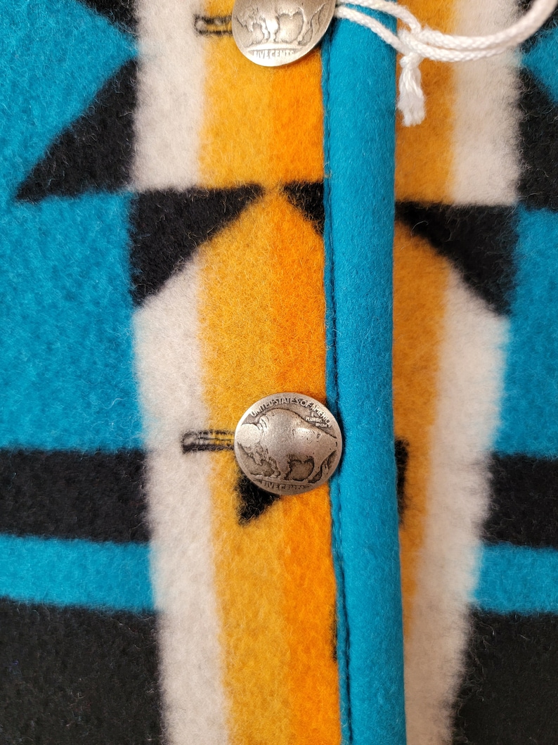 Op de afbeelding: Close-up van een turquoise, zwart, wit en oranje gepatroneerd stof met twee zilveren knopen met een buffelontwerp. De knopen zijn met wit garen aan de stof bevestigd.