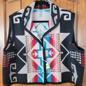 以下が含まれることがあります： 黒、白、赤、青の幾何学模様のベスト。ショールカラーで、柔らかく織られた生地で作られています。ボタン留めで、ハンガーにかけられています。