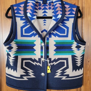 Könnte beinhalten: Eine marineblaue, ärmellose Weste mit einem geometrischen Muster in Blautönen, Grün und Beige. Die Weste hat einen Schalkragen und wird mit zwei silbernen Knöpfen geschlossen.