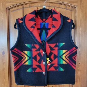 Könnte beinhalten: Eine schwarze Wollweste mit einem roten und schwarzen geometrischen Muster und einem Regenbogenstreifen. Die Weste hat einen Kragen und wird mit silbernen Knöpfen geschlossen.