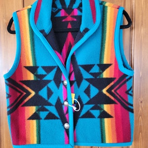 Könnte beinhalten: Eine türkisblaue Weste mit einem bunten geometrischen Muster und einem schwarzen Kragen. Die Weste hat einen Knopfverschluss und ist aus einem weichen, flauschigen Material gefertigt.