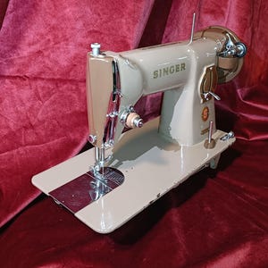 Puede incluir: Máquina de coser Singer vintage en beige y marrón. Cuenta con una aguja plateada, una placa de aguja circular y un gran volante. El logotipo de Singer es visible. La máquina está sobre una base beige. Dimensiones: aproximadamente 40 cm de largo.