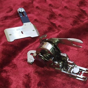 May include: A silver metal sewing machine buttonhole attachment with a dark red adjustment knob and a separate silver metal guide plate featuring a blue accent piece. The attachment has a complex mechanical design with a lever and presser foot.