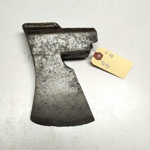 Op de afbeelding: Een verweerde, grijze metalen bijlkop met een licht gebogen blad. De bijlkop mist een deel van de bovenkant. Een geel label met de tekst "F 18 1610" is bevestigd aan de bijlkop.