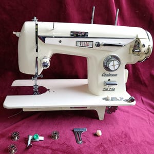 Puede incluir: Máquina de coser Contessa ZIG ZAG vintage de color crema con detalles cromados. La máquina tiene una superficie de trabajo blanca e incluye accesorios, como canillas y un prensatelas. Fondo de color burdeos.