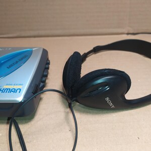May include: A silver and blue Sony Walkman cassette player with the text "WALKMAN" and "WM-EX190" printed on the front. A black pair of Sony headphones with a black cord is next to the Walkman.