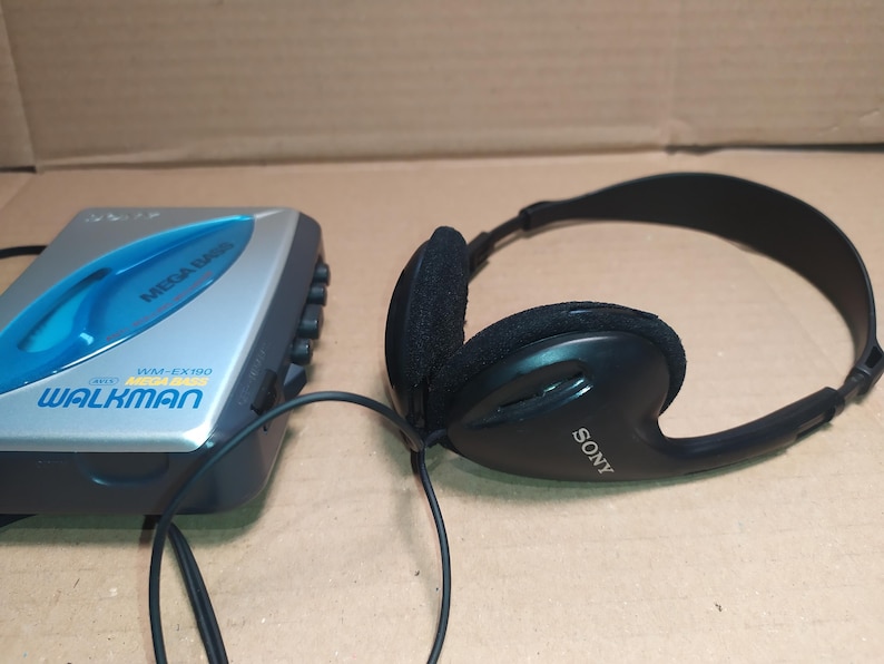 May include: A silver and blue Sony Walkman cassette player with the text "WALKMAN" and "WM-EX190" printed on the front. A black pair of Sony headphones with a black cord is next to the Walkman.