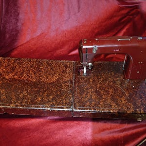 May include: A vintage maroon sewing machine with a matching speckled brown tabletop and a foot pedal. The machine is set against a deep red velvet backdrop, showcasing its classic design and craftsmanship.