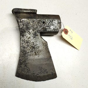 Op de afbeelding: Een roestige metalen bijlkop met een versleten, grijze afwerking. De bijlkop mist de steel en heeft een klein gaatje in de buurt van de bovenkant. Een geel label met de tekst "E4 1337" is aan de bijlkop bevestigd.