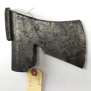 Op de afbeelding: Een verweerde, grijze metalen bijlkop met een licht gebogen blad. De bijlkop is bevestigd aan een houten steel, die niet zichtbaar is op de afbeelding. Een geel label met de tekst "AP8 136 2" is bevestigd aan de bijlkop.