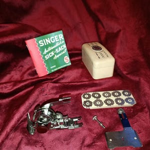 Pode incluir: Um conjunto de acessórios para máquina de costura Singer Automatika Sick-Sack Apparat incluindo manual, caixa de armazenamento creme, calcador de metal, conjunto de discos de cames numerados, um parafuso e uma placa guia azul e prateada.