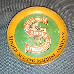 May include: A round vintage metal tin lid featuring a yellow border and a green center. A large red letter S overlays the center, containing the text OMPELUKONEITA SINGER SYMASKINER. An illustration of a person using a sewing machine is visible.