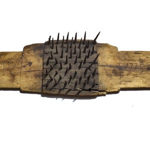 Könnte beinhalten: Ein Holzwerkzeug mit einem Mittelteil, das mit Metallstacheln bedeckt ist. Das Werkzeug hat eine rechteckige Form mit abgerundeten Enden, die jeweils ein kreisförmiges Loch aufweisen. Das Holz hat eine sichtbare Maserung und die Texte "HARD" und "1775" sind sichtbar.