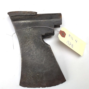 Op de afbeelding: Een verweerde, grijze metalen bijlkop met een gebroken steel. De bijlkop heeft een ruitvormige markering met het nummer "12" erin en de tekst "BILLRKS" erop gestempeld. Een geel label met de tekst "FIL 4 1379" is aan de bijlkop bevestigd.