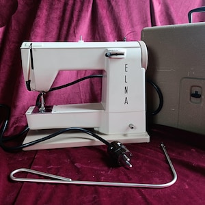 Peut inclure: Machine à coudre Elna blanc cassé vintage avec un cordon d'alimentation noir et un outil en métal argenté. Le nom "ELNA" est imprimé verticalement sur le côté. Une mallette de transport beige est visible en arrière-plan.