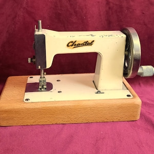 Puede incluir: Máquina de coser Chantal vintage de color crema con base de madera. La máquina presenta un volante y una aguja plateados, con el nombre de la marca en letras doradas. Una herramienta de costura clásica.