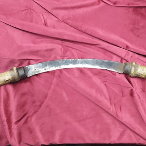 Op de afbeelding: Een gebogen metalen mes met een houten handvat. Het mes is zilverkleurig en het handvat is bruin. Het mes is gebogen en heeft een scherpe punt.