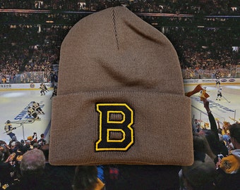 May include: A brown knit beanie with a large yellow "B" embroidered on the front. The beanie is in front of a crowd of people at a hockey game.