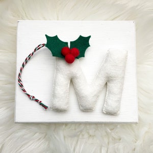 Può includere: Un blocco di legno bianco con la lettera "M" in feltro, decorato con una foglia di agrifoglio verde in feltro e tre bacche rosse in feltro. Un cordino intrecciato rosso, verde e bianco è attaccato. Lo sfondo è una superficie bianca e soffice.