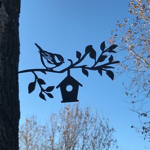 May include: Black metal silhouette of a bird perched on a branch with a birdhouse hanging from the branch. The silhouette is attached to a tree trunk.