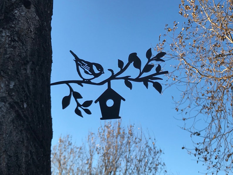 May include: Black metal silhouette of a bird perched on a branch with a birdhouse hanging from the branch. The silhouette is attached to a tree trunk.