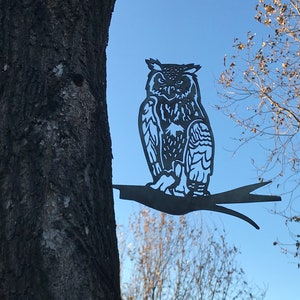 May include: A black metal owl silhouette perched on a branch, mounted on a tree trunk. The owl has detailed feathers and a realistic appearance.
