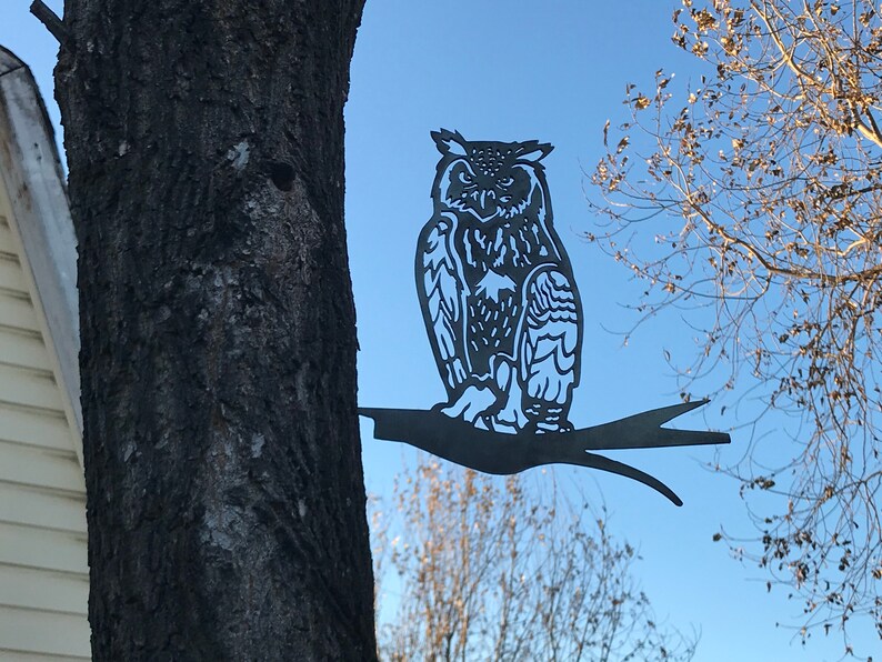 May include: A black metal owl silhouette perched on a branch, mounted on a tree trunk. The owl has detailed feathers and a realistic appearance.
