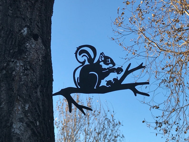 May include: Black metal silhouette of a squirrel sitting on a branch, holding a nut. The squirrel is attached to a tree trunk.