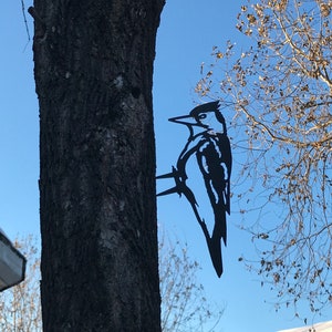 May include: Black metal silhouette of a woodpecker perched on a tree trunk. The silhouette is attached to the tree with a metal bracket. The background is a blue sky with a bare tree branch.
