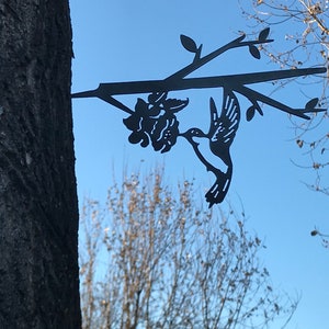 May include: Black metal hummingbird silhouette with a flower attached to a branch, mounted on a tree trunk.