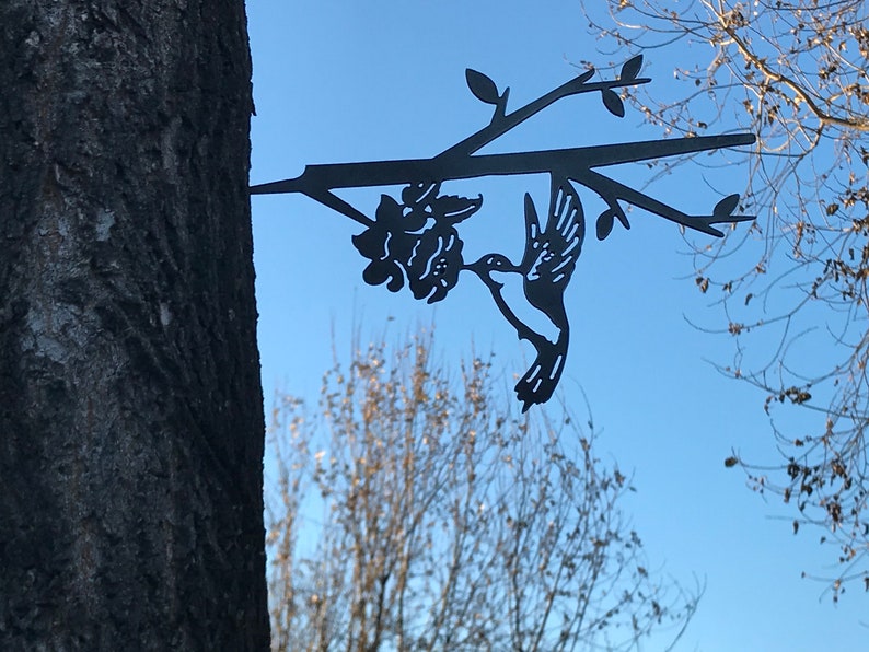 May include: Black metal hummingbird silhouette with a flower attached to a branch, mounted on a tree trunk.