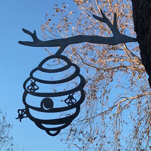 May include: Black metal silhouette of a beehive hanging from a tree branch. The beehive has a round opening and four bees are depicted flying around it.