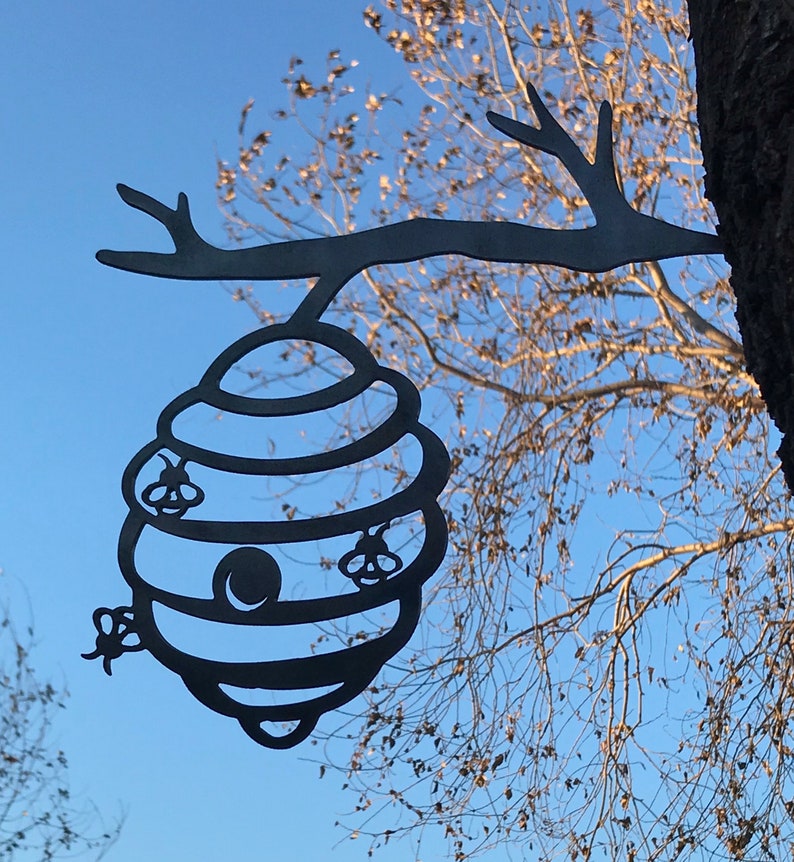 May include: Black metal silhouette of a beehive hanging from a tree branch. The beehive has a round opening and four bees are depicted flying around it.