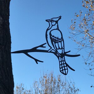 May include: Black metal silhouette of a blue jay perched on a branch. The silhouette is attached to a tree trunk.