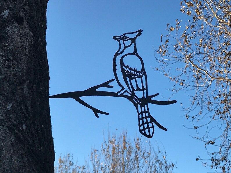 May include: Black metal silhouette of a blue jay perched on a branch. The silhouette is attached to a tree trunk.