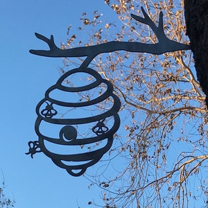May include: A black metal beehive cutout hanging from a tree branch. The beehive has a round opening and three bees are depicted on the sides.