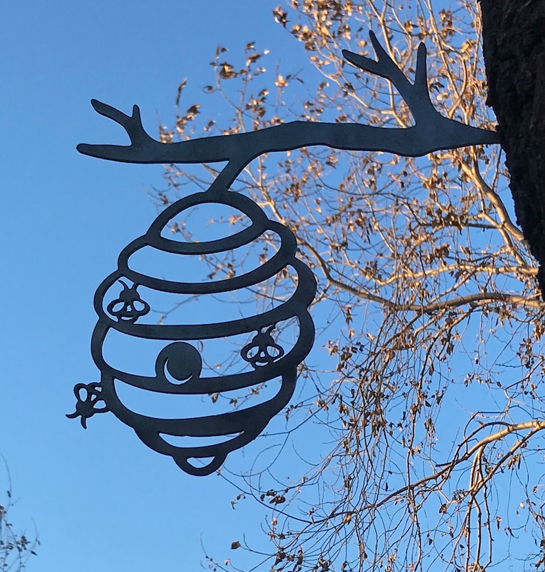 May include: A black metal beehive cutout hanging from a tree branch. The beehive has a round opening and three bees are depicted on the sides.