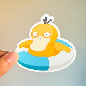 May include: A sticker featuring a cartoon Psyduck character in a blue and white inflatable ring. The Psyduck is yellow with a white beak and black eyes. The sticker is held by a hand.